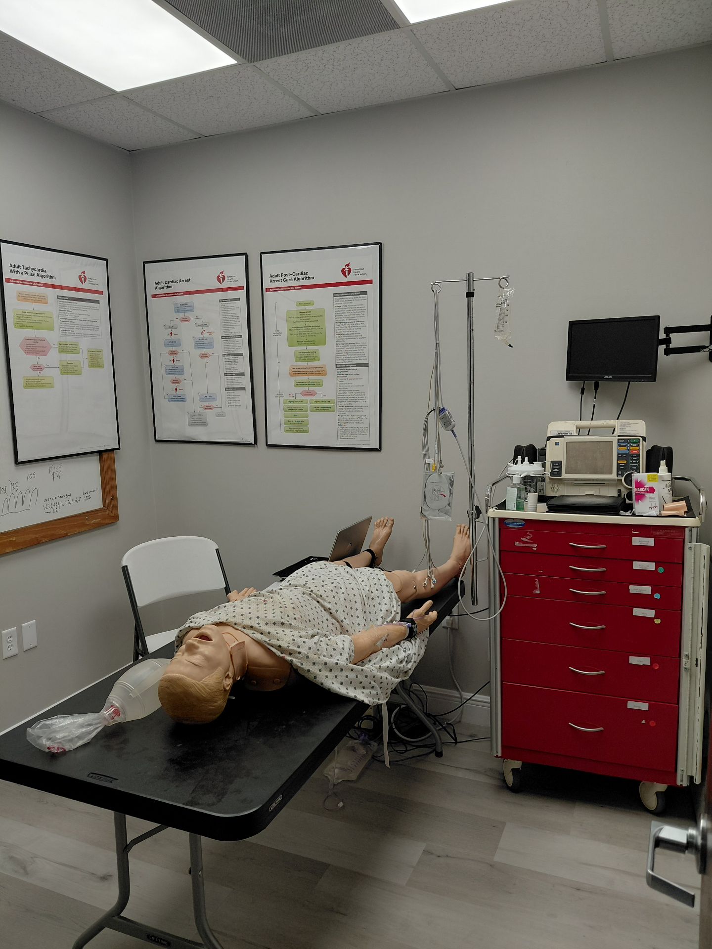 High-fidelity adult manikin with defibrillator, IV pole, and crash cart in ER simulation bay
